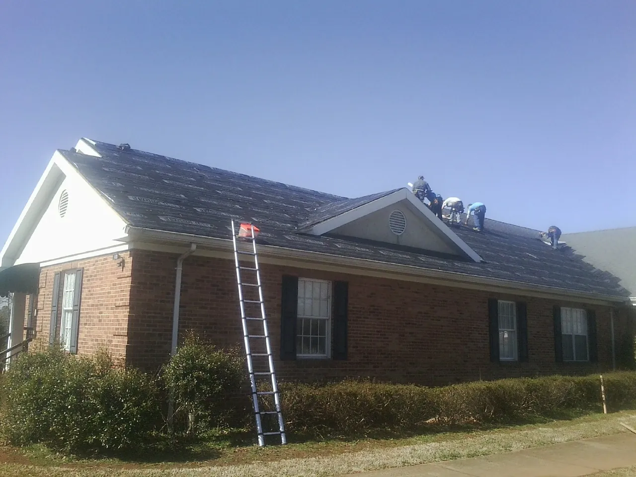 Crew tearing shingles off a brick ranch roof with a ladder set — Metal Roof Repair work by our crew in Colt