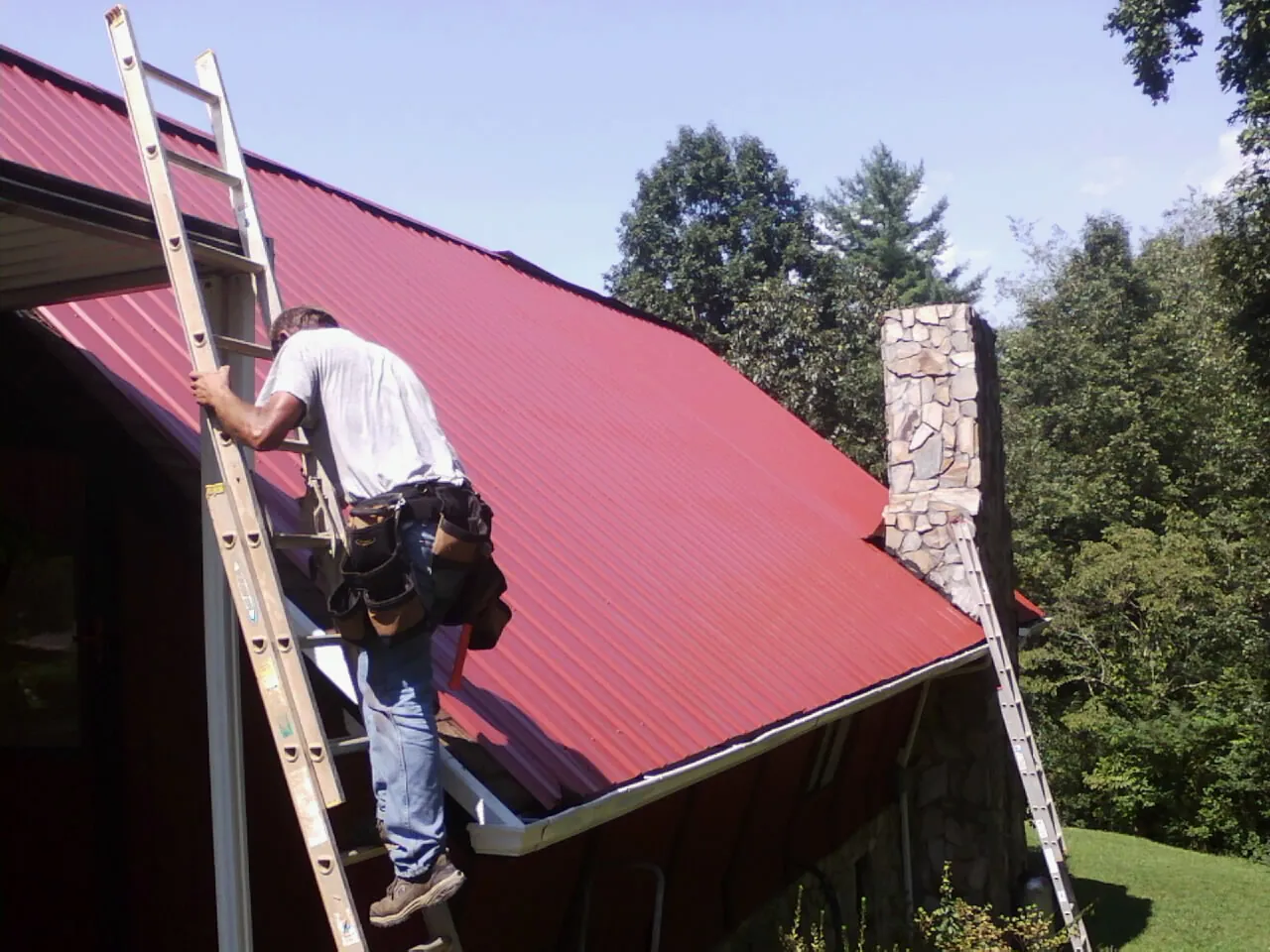 Roofer on a ladder beside a red metal roof and stone chimney — Soffit & Fascia Repair by Roofing Brothers in Colt